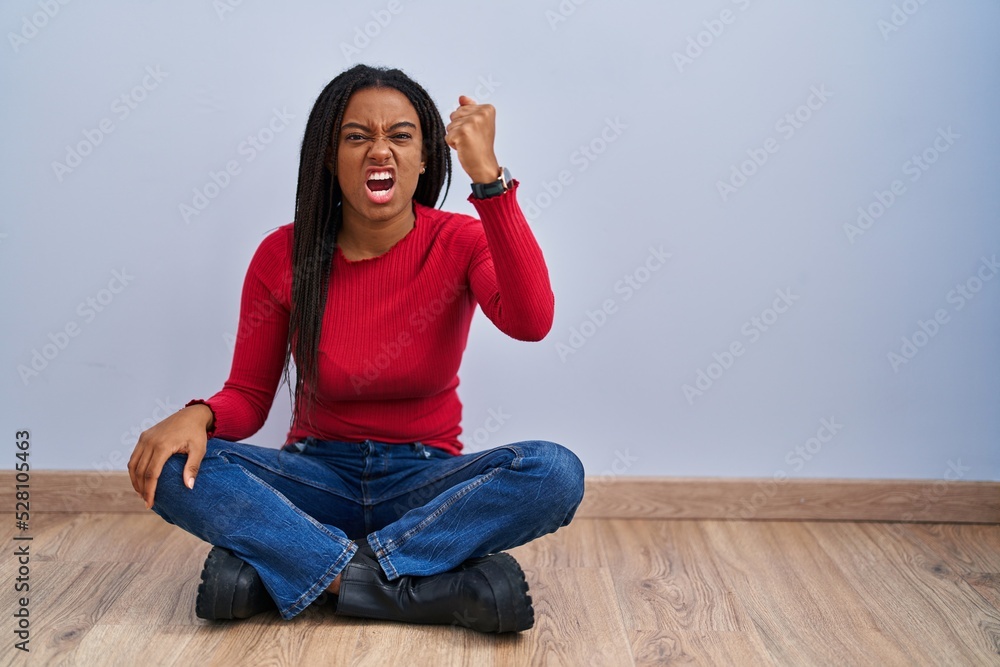 Young african american with braids sitting on the floor at home angry and mad raising fist frustrated and furious while shouting with anger. rage and aggressive concept. Young african american with braids sitting on the floor at home angry and mad raising fist frustrated and furious while shouting with anger. rage and aggressive concept.