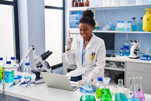 Young african american with braids working at scientist laboratory with laptop smiling with an idea or question pointing finger with happy face  number one