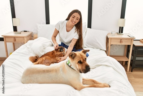 Young hispanic woman playing with dogs sitting on bed at bedroom