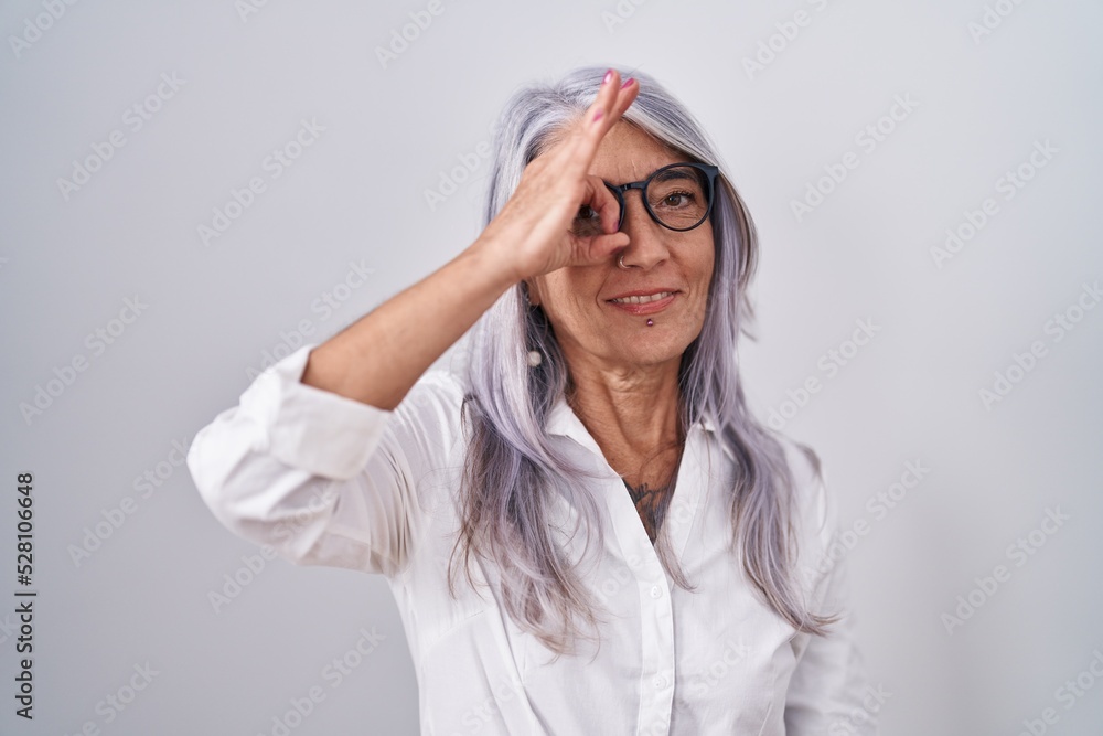 Middle age woman with tattoos wearing glasses standing over white background doing ok gesture with hand smiling, eye looking through fingers with happy face. Middle age woman with tattoos wearing glasses standing over white background doing ok gesture with hand smiling, eye looking through fingers with happy face.
