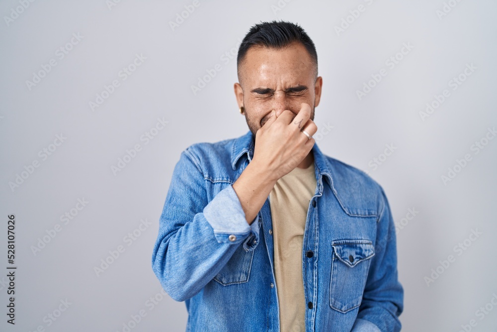 Young hispanic man standing over isolated background smelling something stinky and disgusting, intolerable smell, holding breath with fingers on nose. bad smell Young hispanic man standing over isolated background smelling something stinky and disgusting, intolerable smell, holding breath with fingers on nose. bad smell