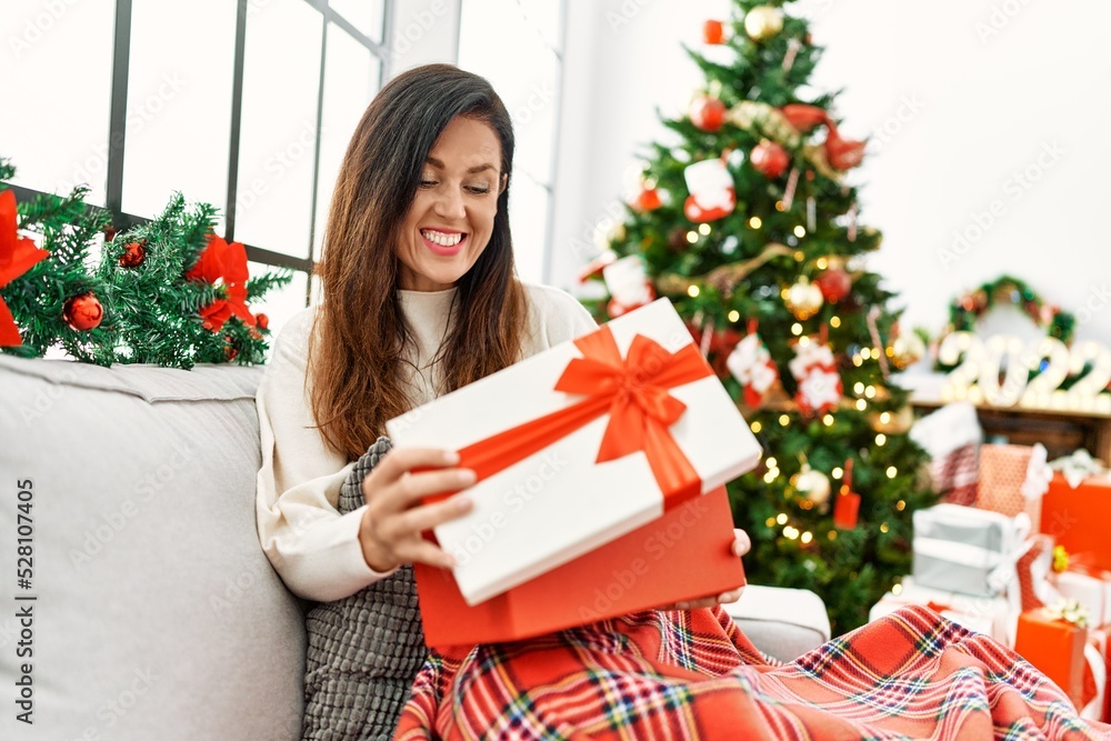 Middle age hispanic woman unboxing gift sitting by christmas tree at home Middle age hispanic woman unboxing gift sitting by christmas tree at home