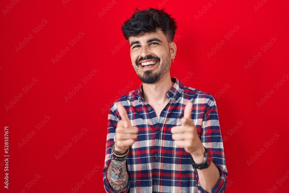 Young hispanic man with beard standing over red background pointing fingers to camera with happy and funny face. good energy and vibes. Young hispanic man with beard standing over red background pointing fingers to camera with happy and funny face. good energy and vibes.