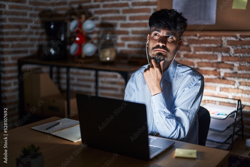 Young hispanic man with beard working at the office at night with hand on chin thinking about question, pensive expression. smiling with thoughtful face. doubt concept. Young hispanic man with beard working at the office at night with hand on chin thinking about question, pensive expression. smiling with thoughtful face. doubt concept.