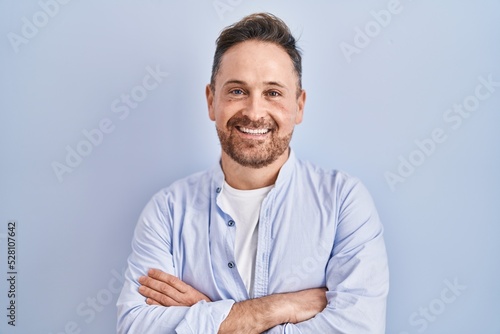 Middle age caucasian man standing over blue background happy face smiling with crossed arms looking at the camera. positive person.