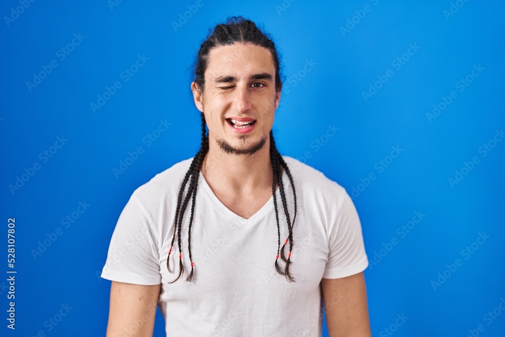 Hispanic man with long hair standing over blue background winking looking at the camera with sexy expression, cheerful and happy face. Hispanic man with long hair standing over blue background winking looking at the camera with sexy expression, cheerful and happy face.