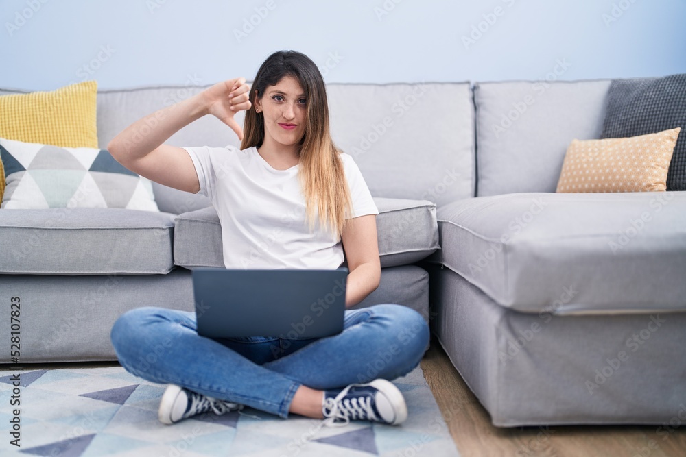 Young brunette woman sitting on the floor at home using laptop with angry face, negative sign showing dislike with thumbs down, rejection concept