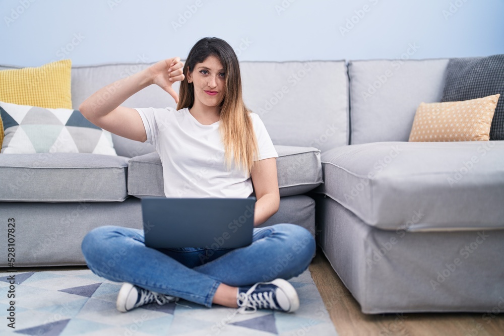 Young brunette woman sitting on the floor at home using laptop with angry face, negative sign showing dislike with thumbs down, rejection concept Young brunette woman sitting on the floor at home using laptop with angry face, negative sign showing dislike with thumbs down, rejection concept