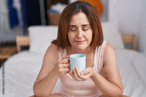 Young woman drinking cup of coffee sitting on bed at bedroom