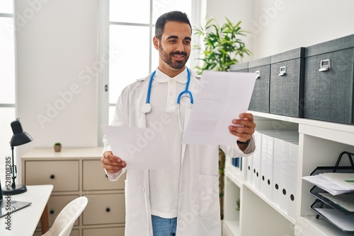 Young hispanic man wearing doctor uniform reading report document at clinic
