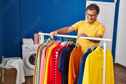 Middle age man smiling confident leaning on clothes rack at laundry room