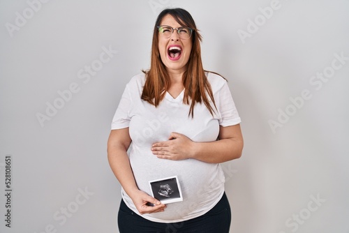 Pregnant woman holding baby ecography angry and mad screaming frustrated and furious, shouting with anger looking up.
