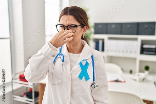 Young brunette doctor woman wearing stethoscope at the clinic smelling something stinky and disgusting, intolerable smell, holding breath with fingers on nose. bad smell