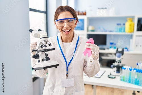 Young brunette woman working at scientist laboratory holding pink ribbon smiling and laughing hard out loud because funny crazy joke.