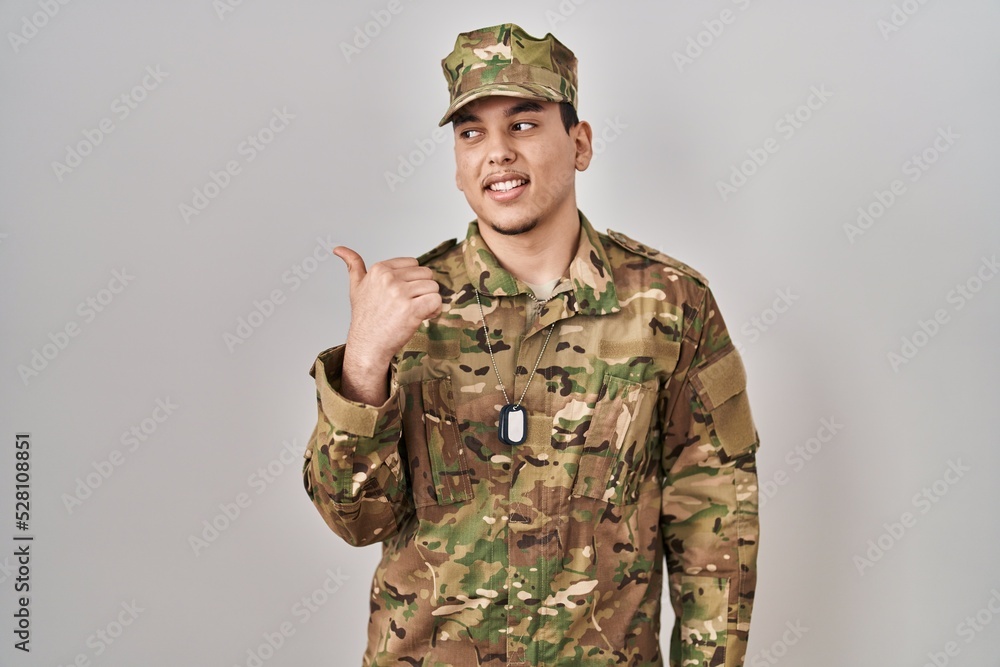 Young arab man wearing camouflage army uniform smiling with happy face looking and pointing to the side with thumb up. Young arab man wearing camouflage army uniform smiling with happy face looking and pointing to the side with thumb up.