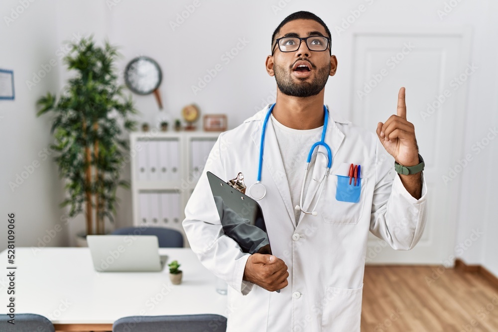 Young indian man wearing doctor uniform and stethoscope amazed and surprised looking up and pointing with fingers and raised arms.