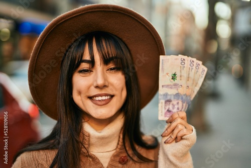 Brunette woman wearing winter hat smiling holding colombian pesos banknotes outdoors at the city