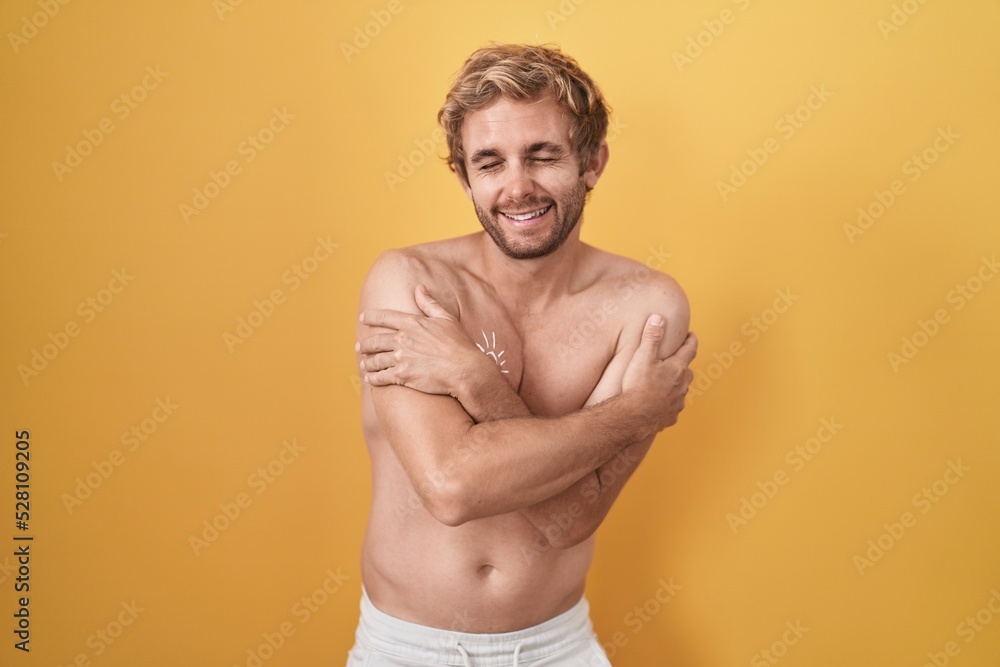 Fototapeta premium Caucasian man standing shirtless wearing sun screen hugging oneself happy and positive, smiling confident. self love and self care