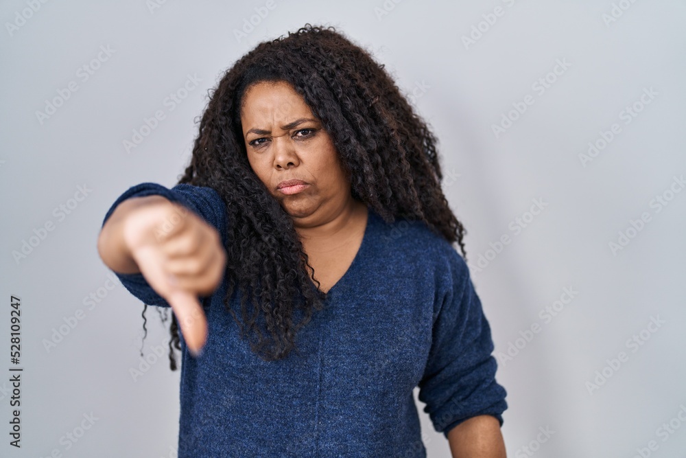 Plus size hispanic woman standing over white background looking unhappy and angry showing rejection and negative with thumbs down gesture. bad expression. Plus size hispanic woman standing over white background looking unhappy and angry showing rejection and negative with thumbs down gesture. bad expression.