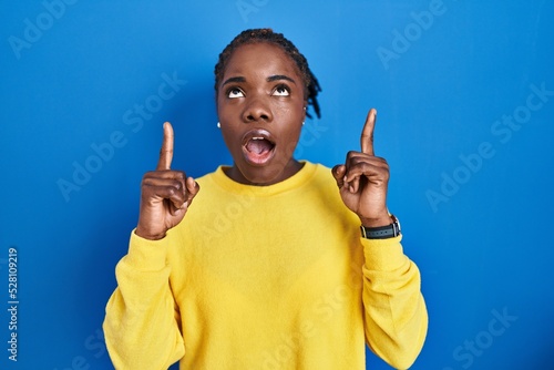 Beautiful black woman standing over blue background amazed and surprised looking up and pointing with fingers and raised arms.