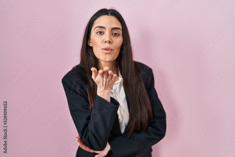 Young brunette woman wearing business style over pink background looking at the camera blowing a kiss with hand on air being lovely and sexy. love expression. Young brunette woman wearing business style over pink background looking at the camera blowing a kiss with hand on air being lovely and sexy. love expression.