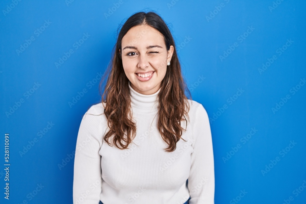 Young hispanic woman standing over blue background winking looking at the camera with sexy expression, cheerful and happy face. Young hispanic woman standing over blue background winking looking at the camera with sexy expression, cheerful and happy face.