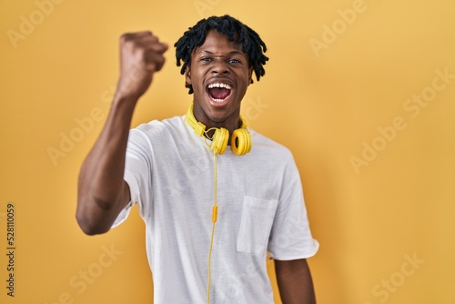 Young african man with dreadlocks standing over yellow background angry and mad raising fist frustrated and furious while shouting with anger. rage and aggressive concept.