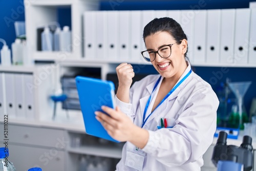 Young brunette woman working at scientist laboratory using tablet screaming proud  celebrating victory and success very excited with raised arm
