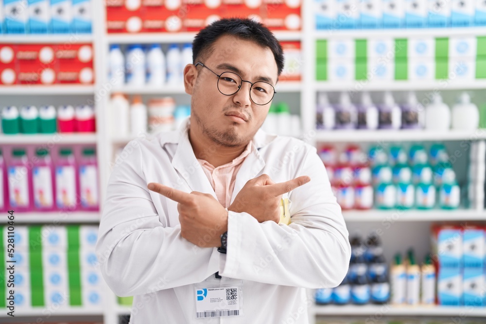 Chinese young man working at pharmacy drugstore pointing to both sides with fingers, different direction disagree Chinese young man working at pharmacy drugstore pointing to both sides with fingers, different direction disagree