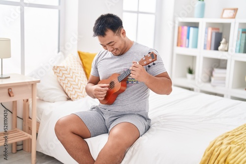 Young chinese man playing ukulele sitting on bed at bedroom