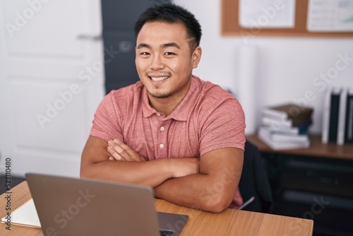 Young chinese man business worker using laptop sitting with arms crossed gesture at office
