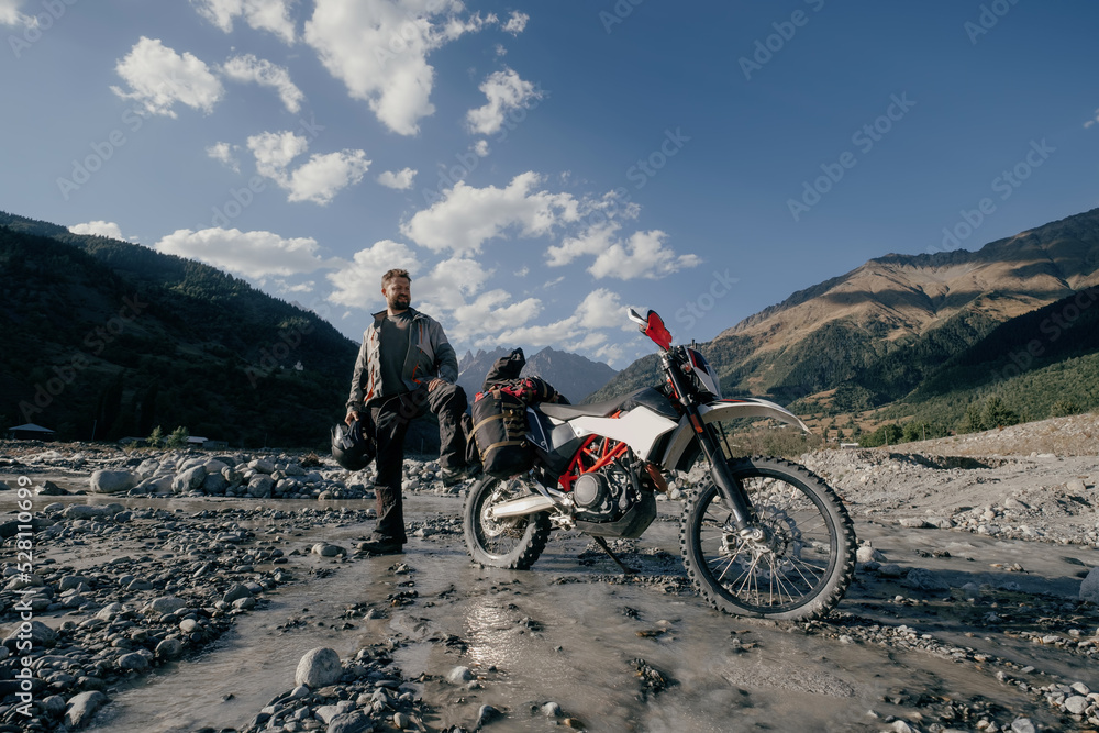 Happy biker Man wearing beard and motorcycle clothes in long moto trip standing in mountains on river near his motorcycle
