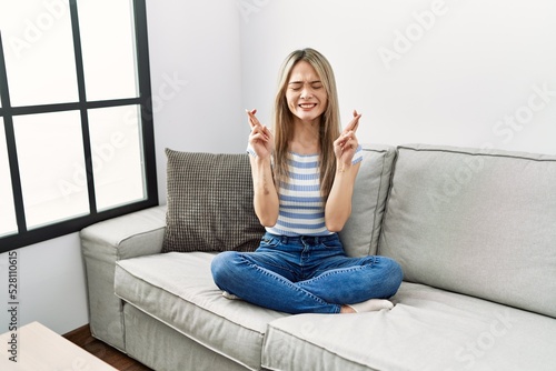 Asian young woman sitting on the sofa at home gesturing finger crossed smiling with hope and eyes closed. luck and superstitious concept.