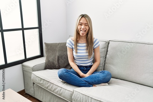 Asian young woman sitting on the sofa at home sticking tongue out happy with funny expression. emotion concept.