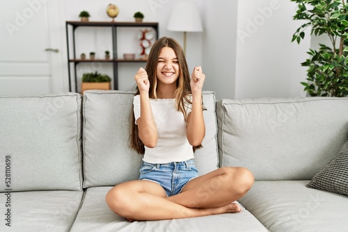 Young brunette teenager sitting on the sofa at home excited for success with arms raised and eyes closed celebrating victory smiling. winner concept.
