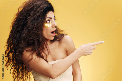 Shocked curly pretty Latin female with gold hydrogel patches under eye point finger look aside posing isolated over yellow background. Cosmetic product ad Natural beauty concept Studio profile
