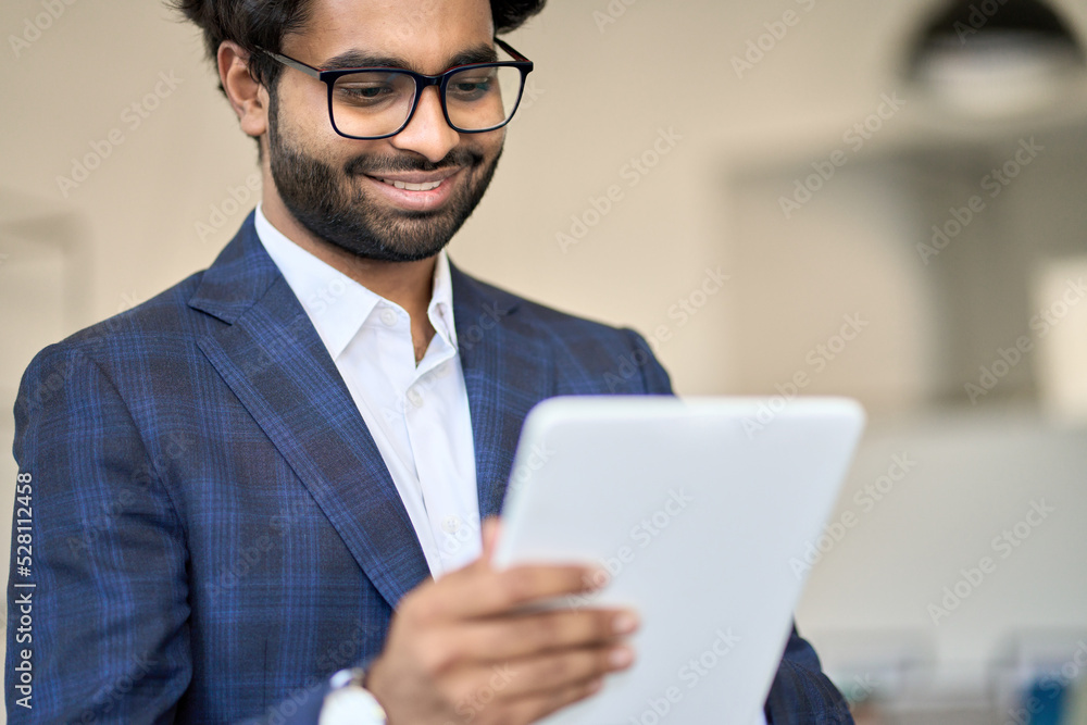 Smiling elegant young indian business man professional project manager, arab company employee, eastern businessman executive wearing suit and glasses standing in office holding using digital tablet. Smiling elegant young indian business man professional project manager, arab company employee, eastern businessman executive wearing suit and glasses standing in office holding using digital tablet.