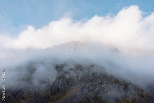 Lovely scenery with big mountain peak in thick clouds at early morning. Scenic view to high pinnacle in gantly blue cloudy sky in sunlight. Large mountain top in low clouds. Sharp rocks in dense fog.
