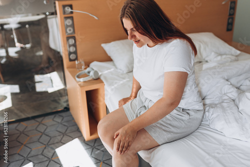 Young woman sitting on bed and feeling knee pain, she massage her knee at home. Healthcare and medical concept. Pain in the knee.