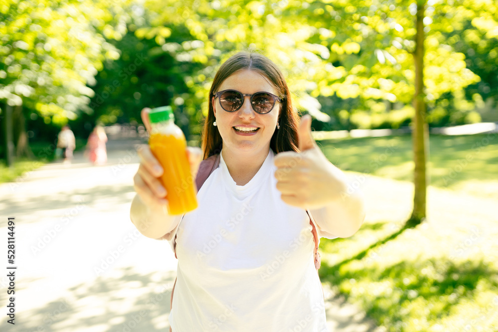 Young stylish happy woman with juice giving thumb up as sign of success and standing over nature background. Redhead girl walking in the park and holding a bottle of fresh cold smoothie in a sunny day Young stylish happy woman with juice giving thumb up as sign of success and standing over nature background. Redhead girl walking in the park and holding a bottle of fresh cold smoothie in a sunny day