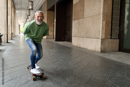 Active energetic happy cool gray haired old adult man skateboarding on city street, smiling sporty fit middle aged mature older male professional skater having fun enjoying riding skateboard outdoor.