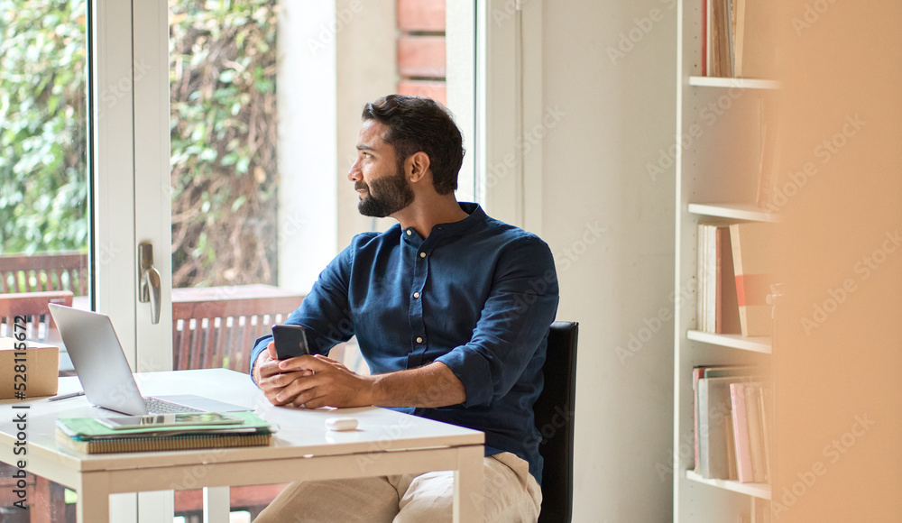 Fototapeta premium Authentic shot of indian professional business man, thoughtful eastern businessman, ethnic entrepreneur holding smartphone online remote working at home in office sitting at desk looking at window.