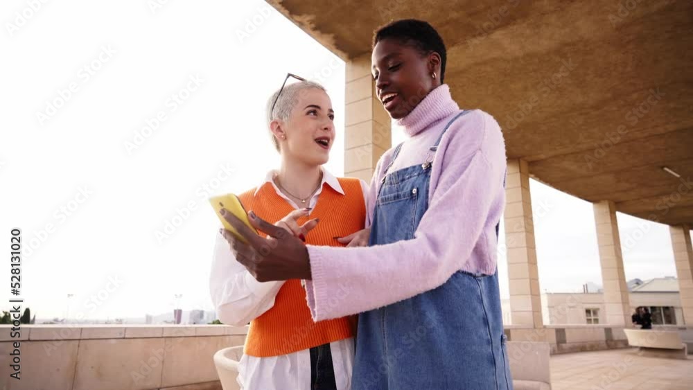 Two cheerful women friends using mobile cell. Happy African American and caucasian girl shaved head having fun outdoors watching something at smart phone and smiling