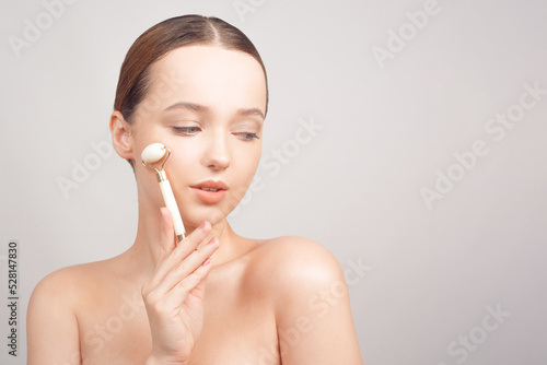 Beauty face care. Woman doing face massage with white jade facial rollers for spa skin care treatment at home. Girl model using natural massager tool portrait on white .