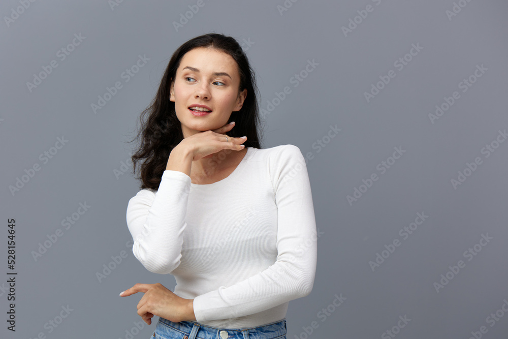 Obraz premium a sophisticated woman stands on a gray background in a white T-shirt, looks mysteriously to the side, touching her chin with her hand, putting her other hand in front of her