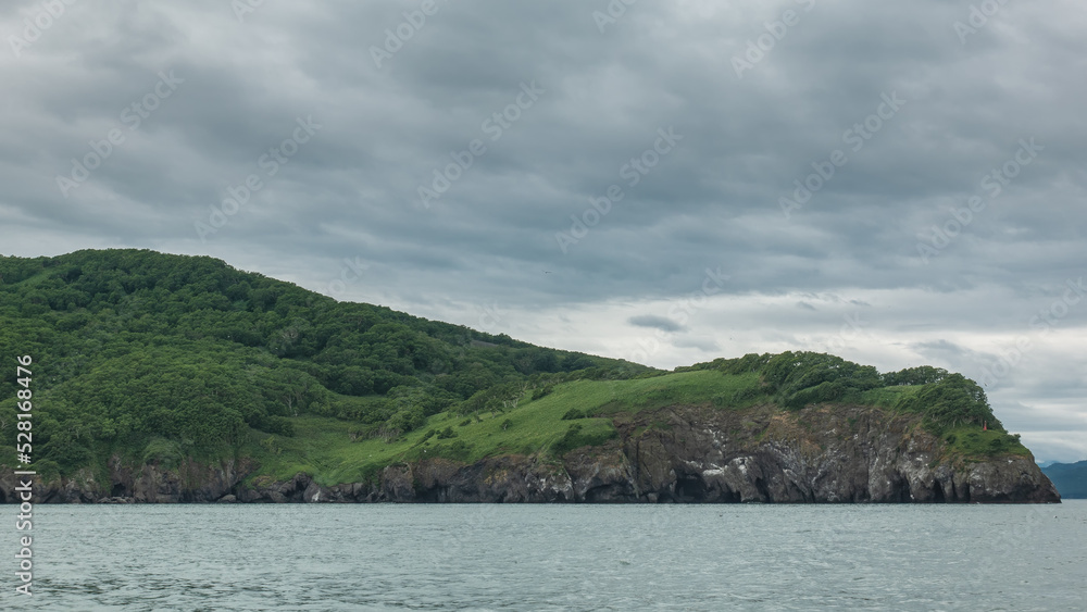 The picturesque coast of Kamchatka against the background of cloudy sky and ocean.  Green hills and steep cliffs. Avacha Bay