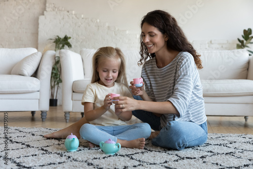 Foto Little cute daughter play with Latina mother at home, holding plastic toy dishware, imagining tea-party seated on warm floor in living room