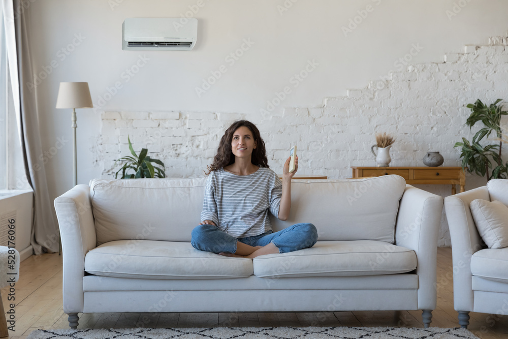 Attractive Hispanic woman holds remote controller turns on AC set comfort temperature in modern living room, enjoy fresh conditioned air inside. Climate control at home for comfortable life concept Attractive Hispanic woman holds remote controller turns on AC set comfort temperature in modern living room, enjoy fresh conditioned air inside. Climate control at home for comfortable life concept