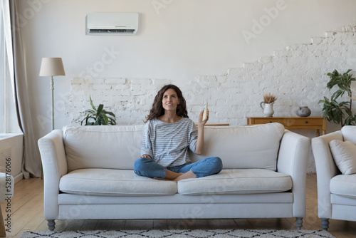 Attractive Hispanic woman holds remote controller turns on AC set comfort temperature in modern living room, enjoy fresh conditioned air inside. Climate control at home for comfortable life concept