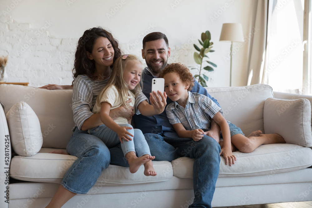 Happy parents with little 5s children spend leisure using smartphone. Couple and siblings have fun enjoy mobile app, make video call, take selfie picture for memory. Pastime of family and tech concept